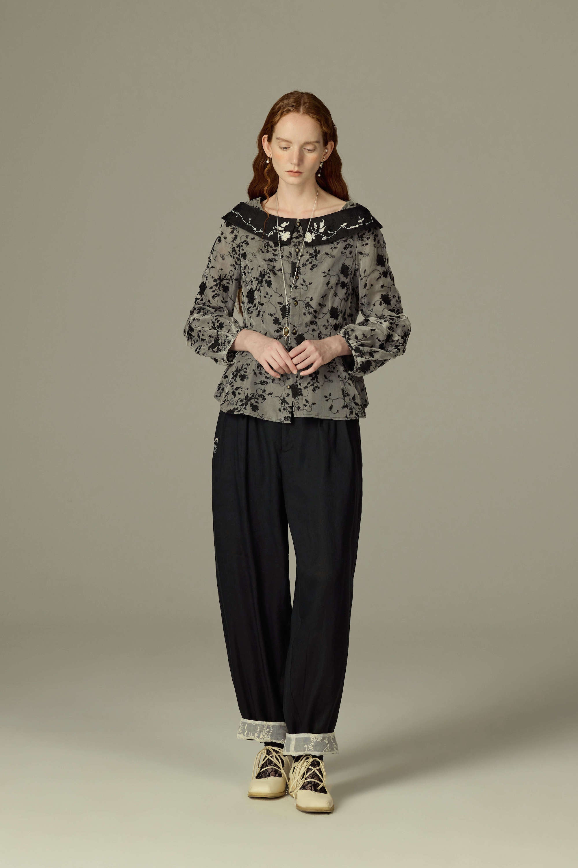 A woman modeling a floral print top and black trousers, standing against a gray background.